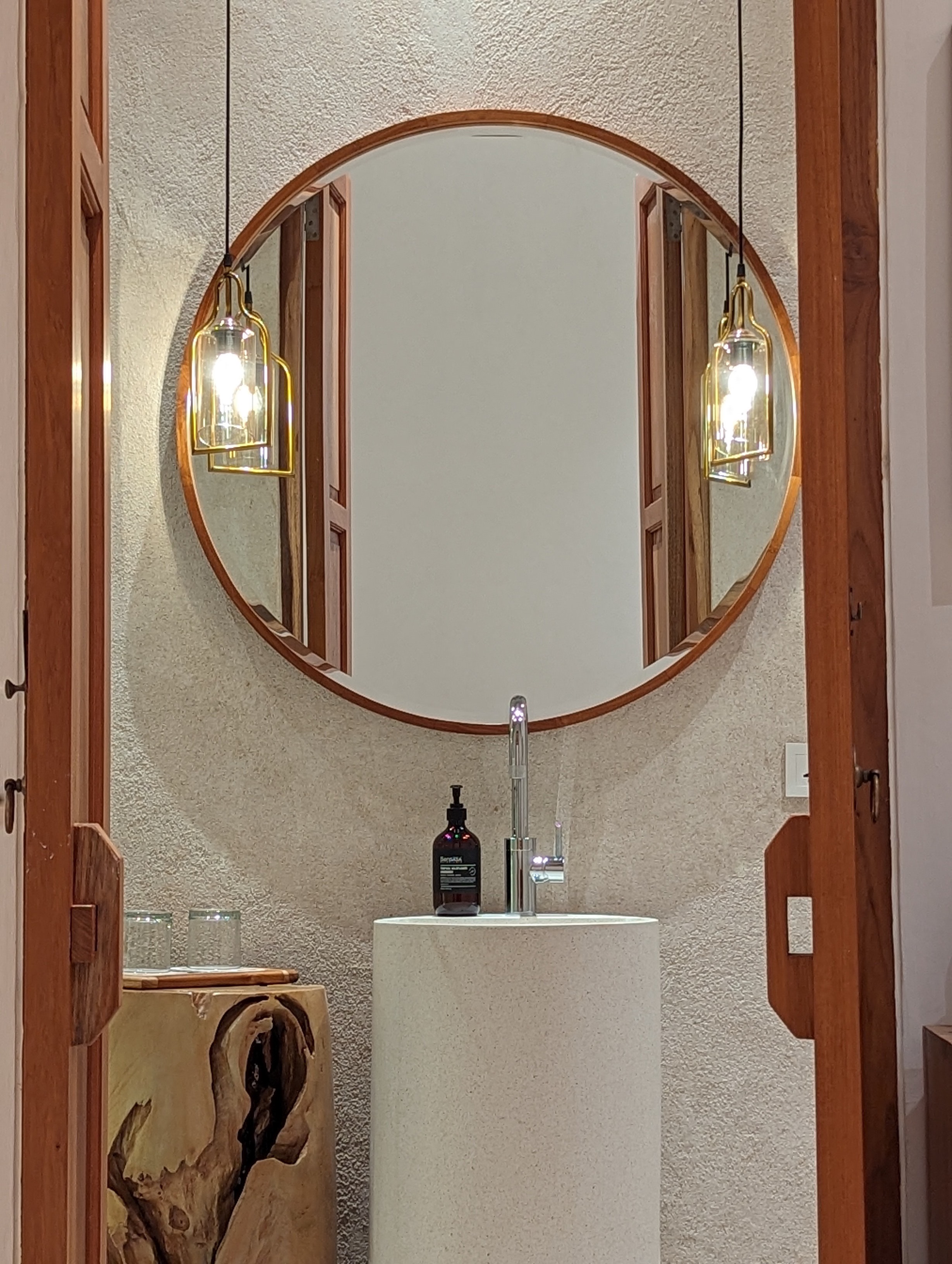 Round teak-framed mirror and brass pendant lamps above a stone pedestal sink